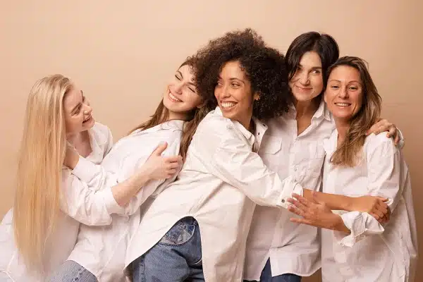 Mulheres diferentes e bonitas, brincam e sorriem felizes umas com as outras. Todas vestem camisa branca e calça jeans.