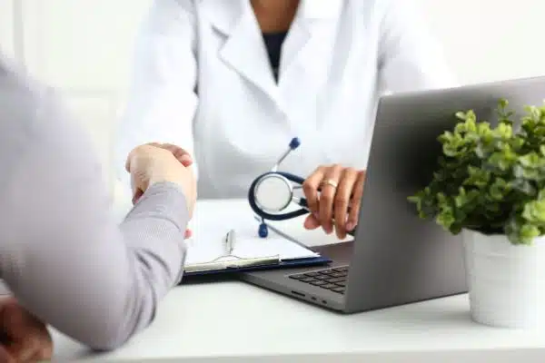 Médica segura um estetoscópio na mão esquerda, frente ao notebook, e cumprimenta paciente com a mão direita.