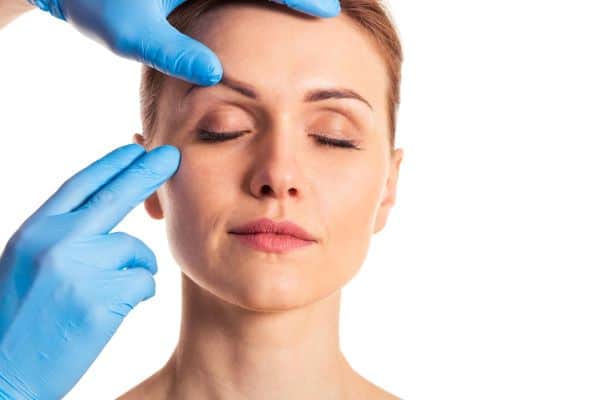 Rosto de uma mulher jovem sendo analisado por médico que veste luvas azuis para a realização de uma blefaroplastia não cirúrgica.