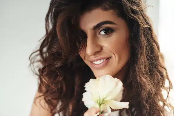 Linda mulher com cabelos escuros, compridos eencaracolados, sorri segurando uma flor branca próxima ao queixo.
